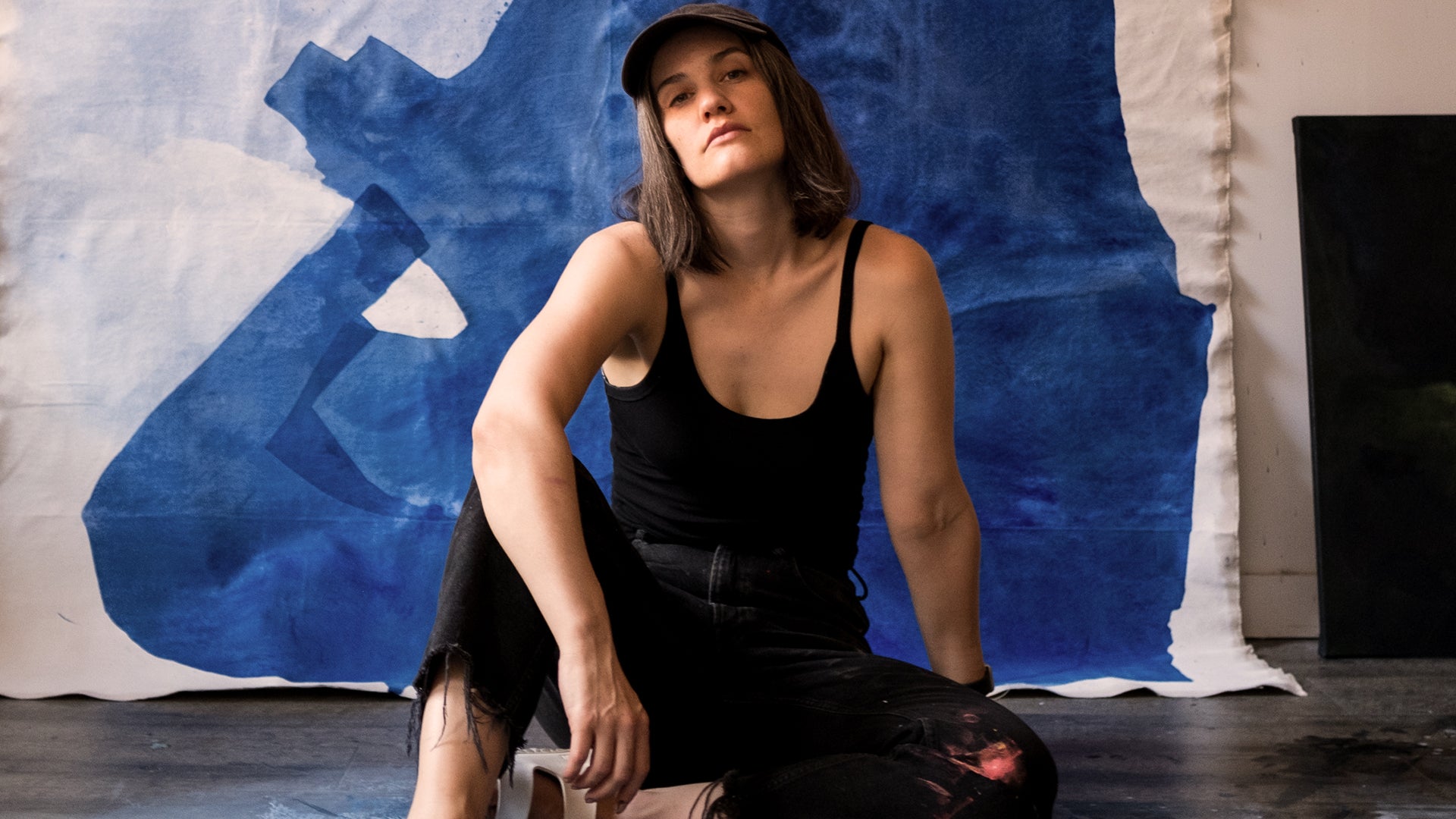 Image of the artist sitting on the ground in front of one of her unfinished paintings on canvas, staring into the camera