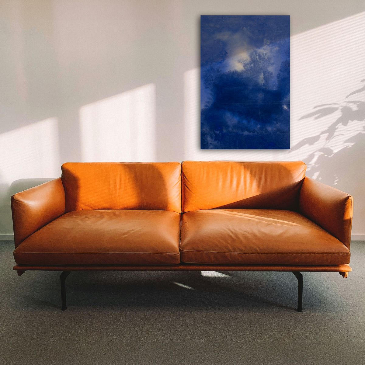 A blue abstract painting hangs above a brown leather coach in a sunny room