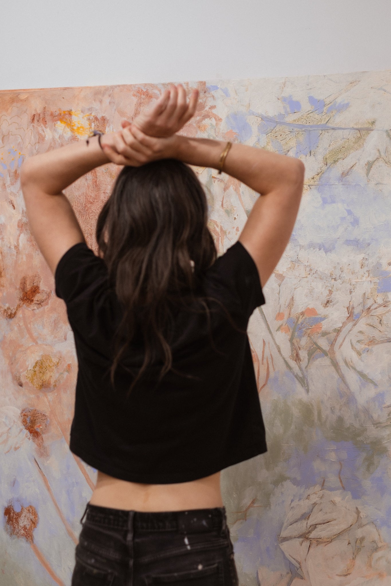 Person wearing a black crop top and dark pants with arms raised in front of a colorful abstract painting.